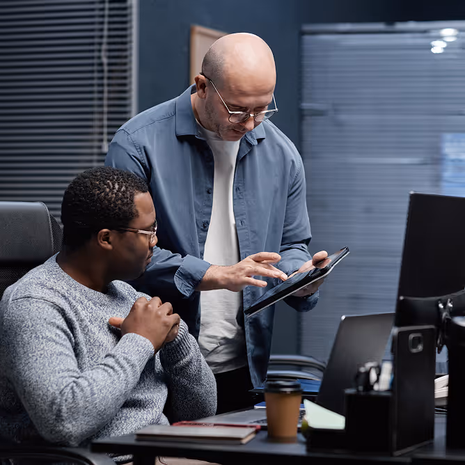 Two men in an office, one sitting and watching while the other stands and works on a tablet.