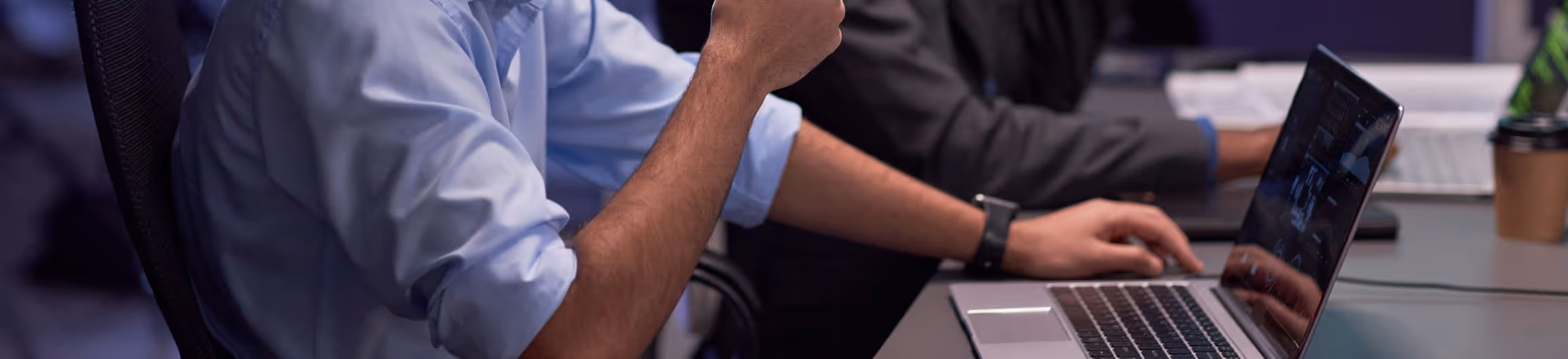 Two people working on laptops at a desk with one person wearing a light blue shirt and the other in a dark jacket.